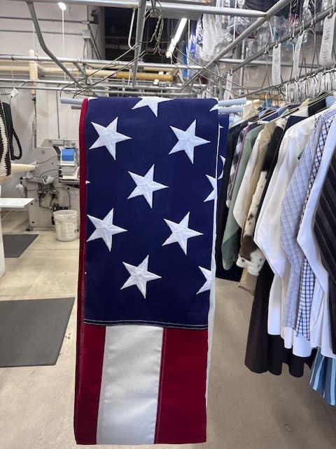 A folded American flag hangs on a rack inside a dry cleaning shop, surrounded by other cleaned and pressed garments.