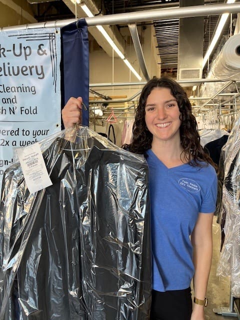 Woman in a blue shirt stands in a dry cleaning facility, holding a freshly cleaned and bagged suit.