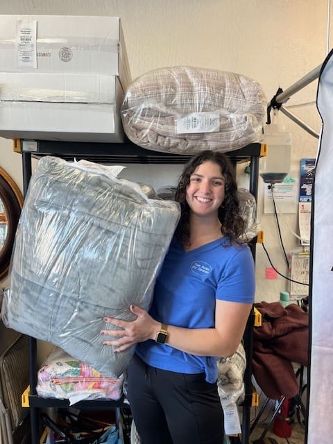 A woman wearing a blue shirt holds a large, packaged gray blanket while standing in front of a shelving unit filled with other blankets and boxes.