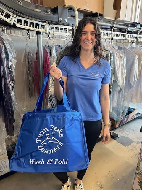 A woman in a blue shirt stands in a dry cleaning shop, holding a blue "Twin Peaks Cleaners Wash & Fold" bag. Garments on hangers are visible in the background.