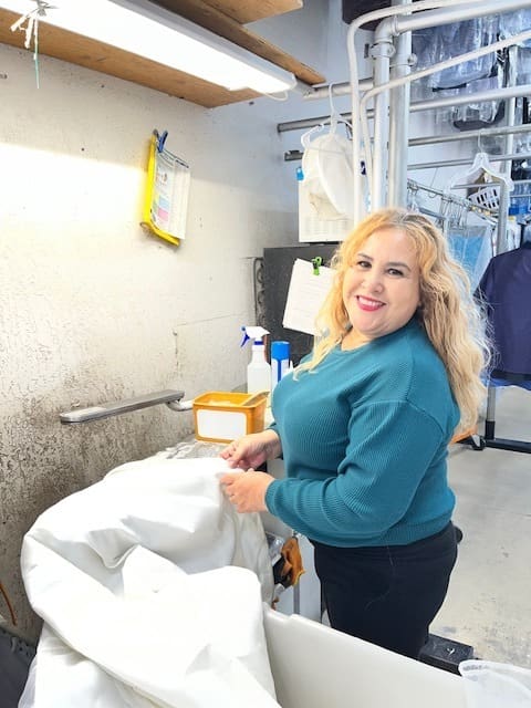 A woman with blonde hair and a teal sweater stands next to laundry equipment, handling white fabric in a dry cleaning or laundry facility.