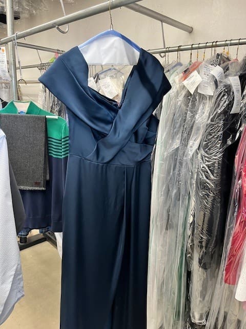 A dark blue formal dress with a draped neckline hangs on a rack among other clothes, some covered in plastic garment bags.