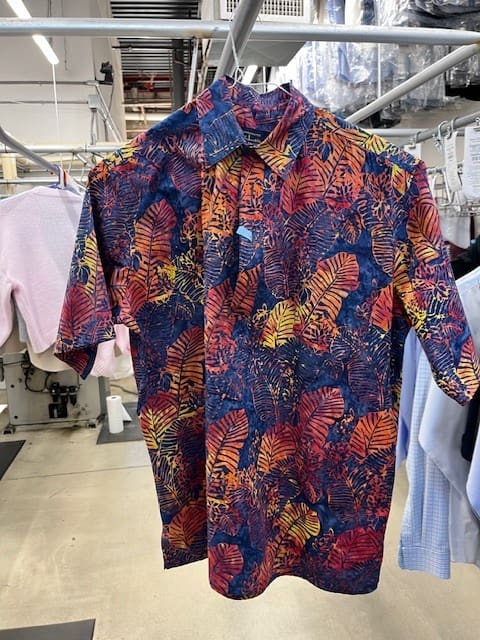 A brightly colored short-sleeve button-up shirt with a tropical leaf pattern hangs on a clothing rack in a laundry or dry cleaning facility.