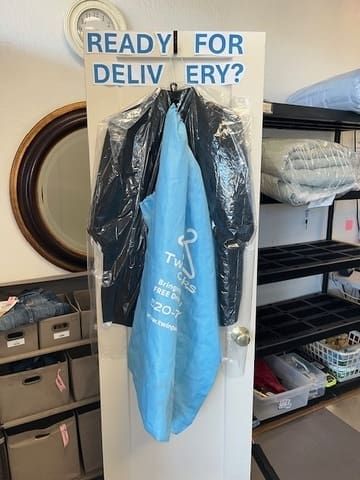 A dry cleaning bag and clothes hang on a door labeled "READY FOR DELIVERY?" in a laundry or dry cleaning shop with storage bins and shelves nearby.
